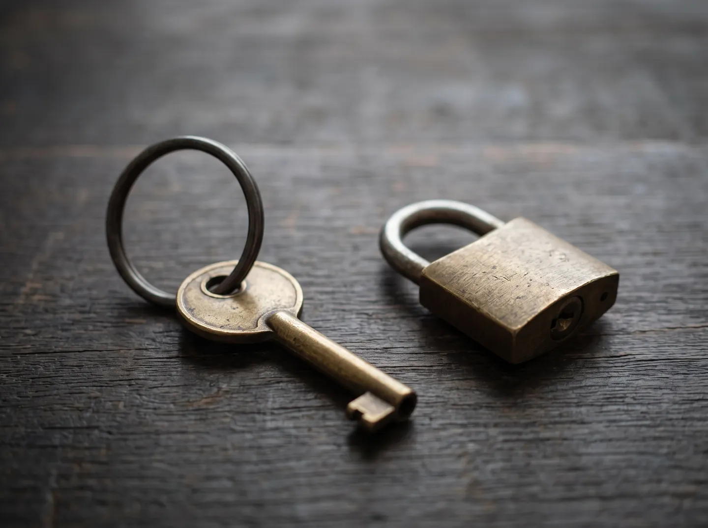A single old brass key on a ring resting on a dark wooden desk beside a small padlock